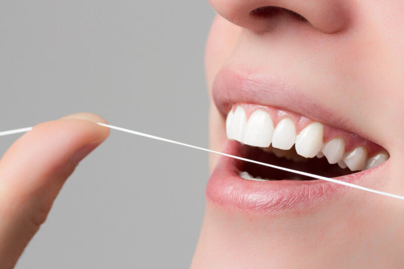 Una persona está usando hilo dental frente a un espejo de baño bien iluminado, aplicando la técnica adecuada para limpiar entre los dientes y la línea de las encías. Este hábito de higiene bucal es fundamental para eliminar restos de comida y prevenir problemas como caries y gingivitis.