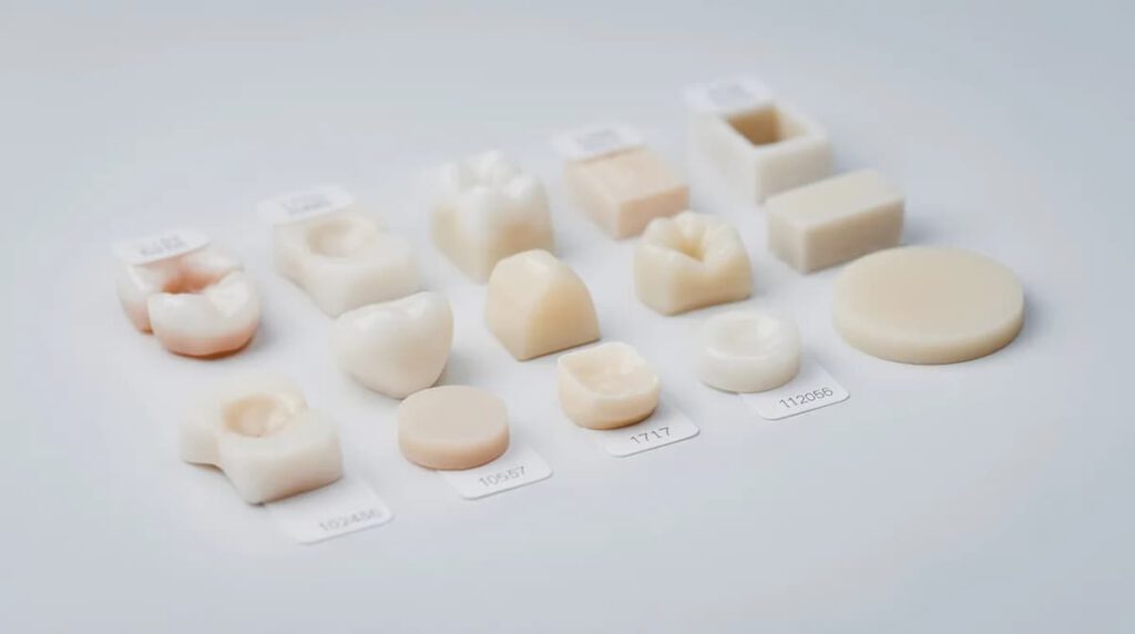 Muestras de diferentes materiales cerámicos para coronas dentales, incluyendo porcelana y zirconio, están expuestas sobre una superficie blanca de laboratorio, mostrando su variedad en color y textura. Estas piezas son fundamentales en tratamientos odontológicos para restaurar dientes dañados y mejorar la estética de la sonrisa del paciente.