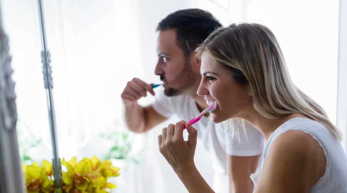 A smiling couple brush their teeth together in a bright bathroom, promoting good oral hygiene habits and the importance of brushing for oral health. The cheerful atmosphere highlights the daily tooth-brushing routine, which is essential for preventing problems such as tooth decay and gum disease.