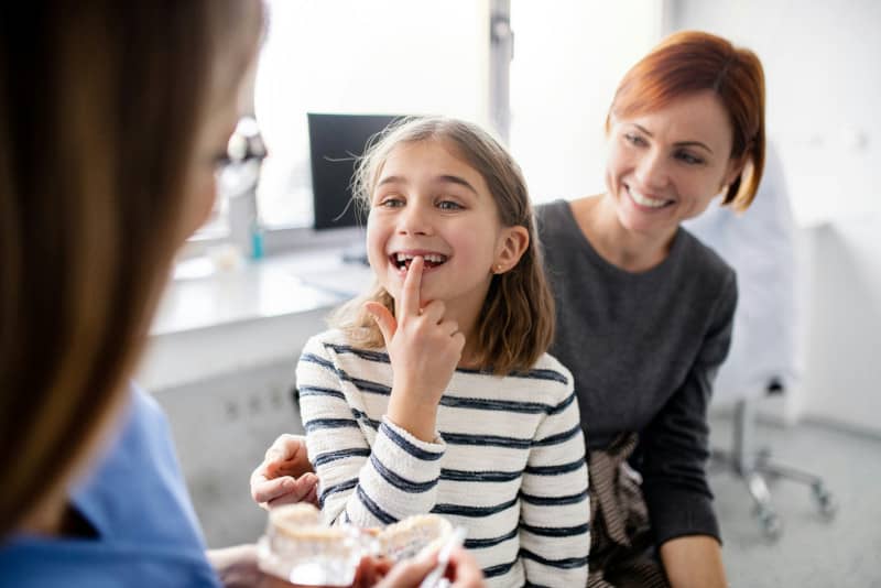 Una niña sonriente muestra sus dientes durante una revisión dental, destacando su sonrisa y la importancia de la salud bucal. Esta imagen ilustra la consulta con el dentista, donde se pueden identificar problemas como dientes torcidos en niños y la necesidad de un tratamiento ortodóntico.