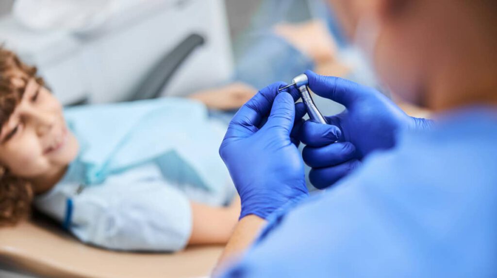 Un niño sonriente está sentado en un sillón dental, mientras el dentista le muestra un espejo para que vea sus dientes. La escena refleja la importancia de la salud dental y la corrección de problemas como los dientes torcidos en niños.